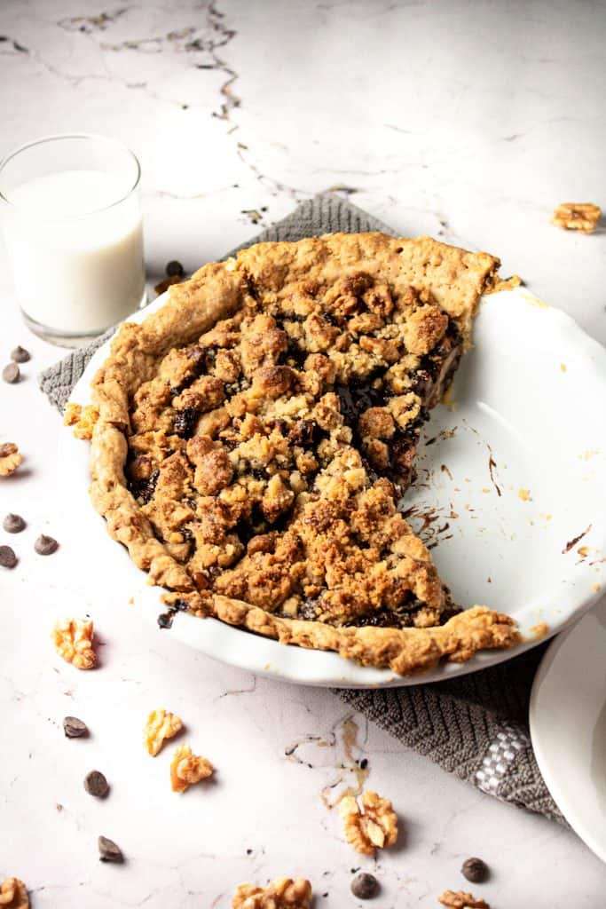 Up close view of a pear chocolate crumble pie with a slice missing, a glass of milk in the background and walnuts and chocolate chips in the foreground