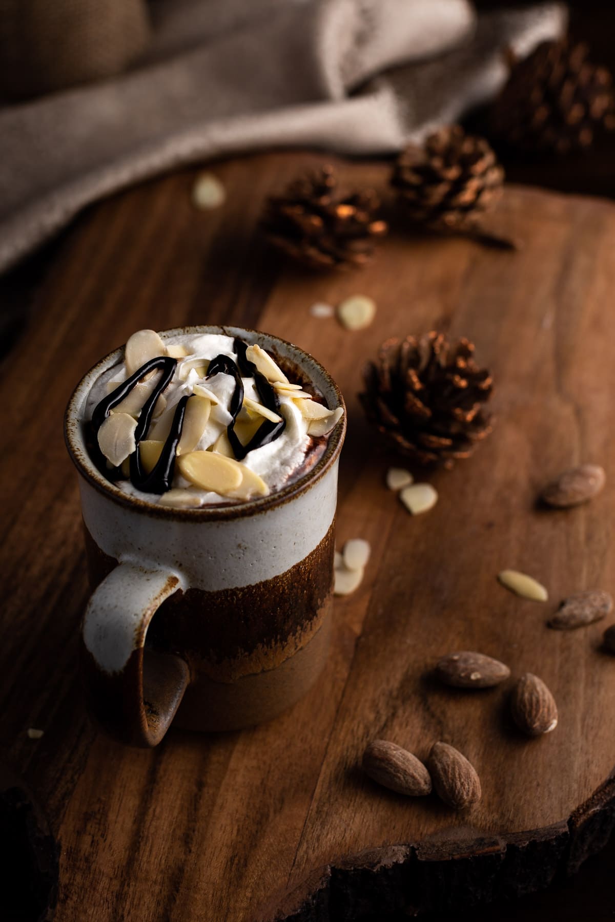 A brown mug of hot chocolate topped with whipped cream and almonds, next to scattered almonds.