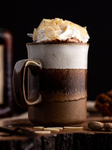 Side view of a brown mug of dark hot chocolate with amaretto, topped with whipped cream and sliced almonds, in front of a black background.