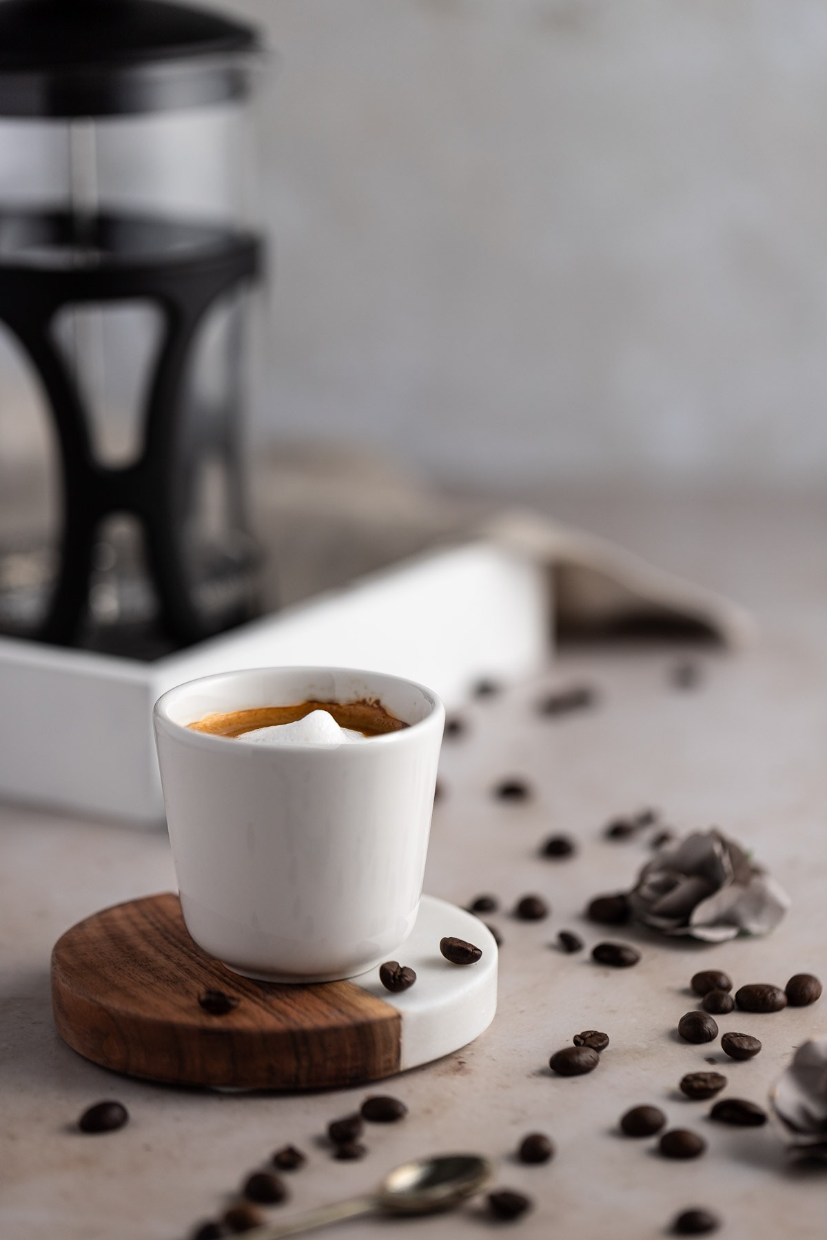 A traditional espresso macchiato in a small white espresso mug on a white and wooden coaster, surrounded by scattered coffee beans.