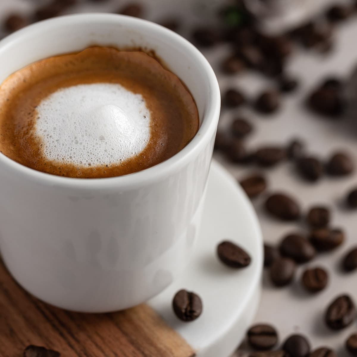 A small white espresso mug filled with an espresso macchiato, surrounded by scattered coffee beans. 