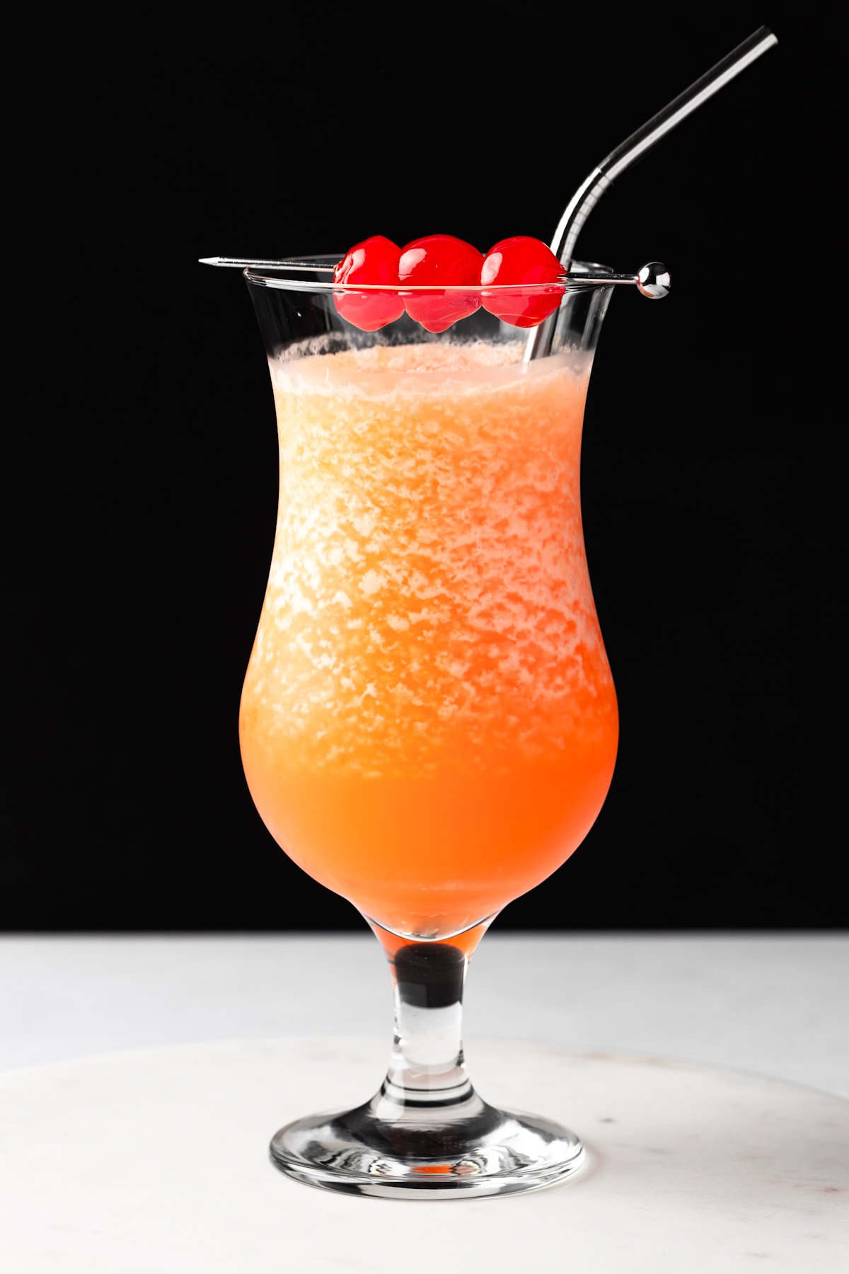 An orange frozen hurricane cocktail garnished with maraschino cherries on a black background.