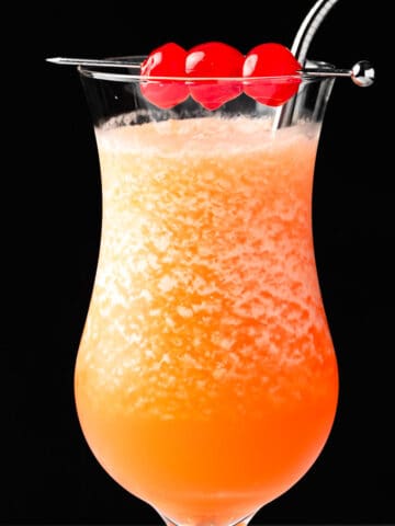An orange frozen hurricane cocktail garnished with maraschino cherries on a black background.