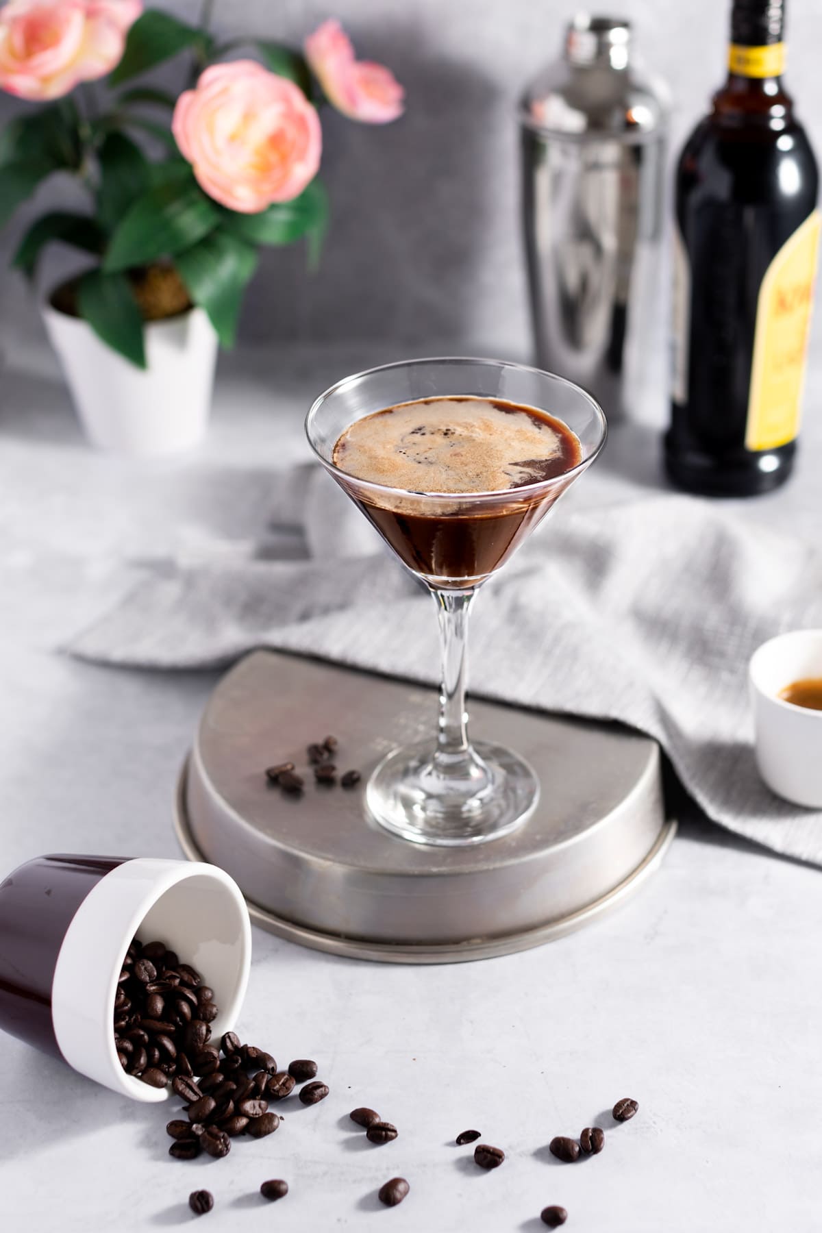 A martini sitting on a flipped over round metal tin, next to a cup of spilled coffee beans, with pink flowers, a cocktail shaker and bottle of kahlua in the background.