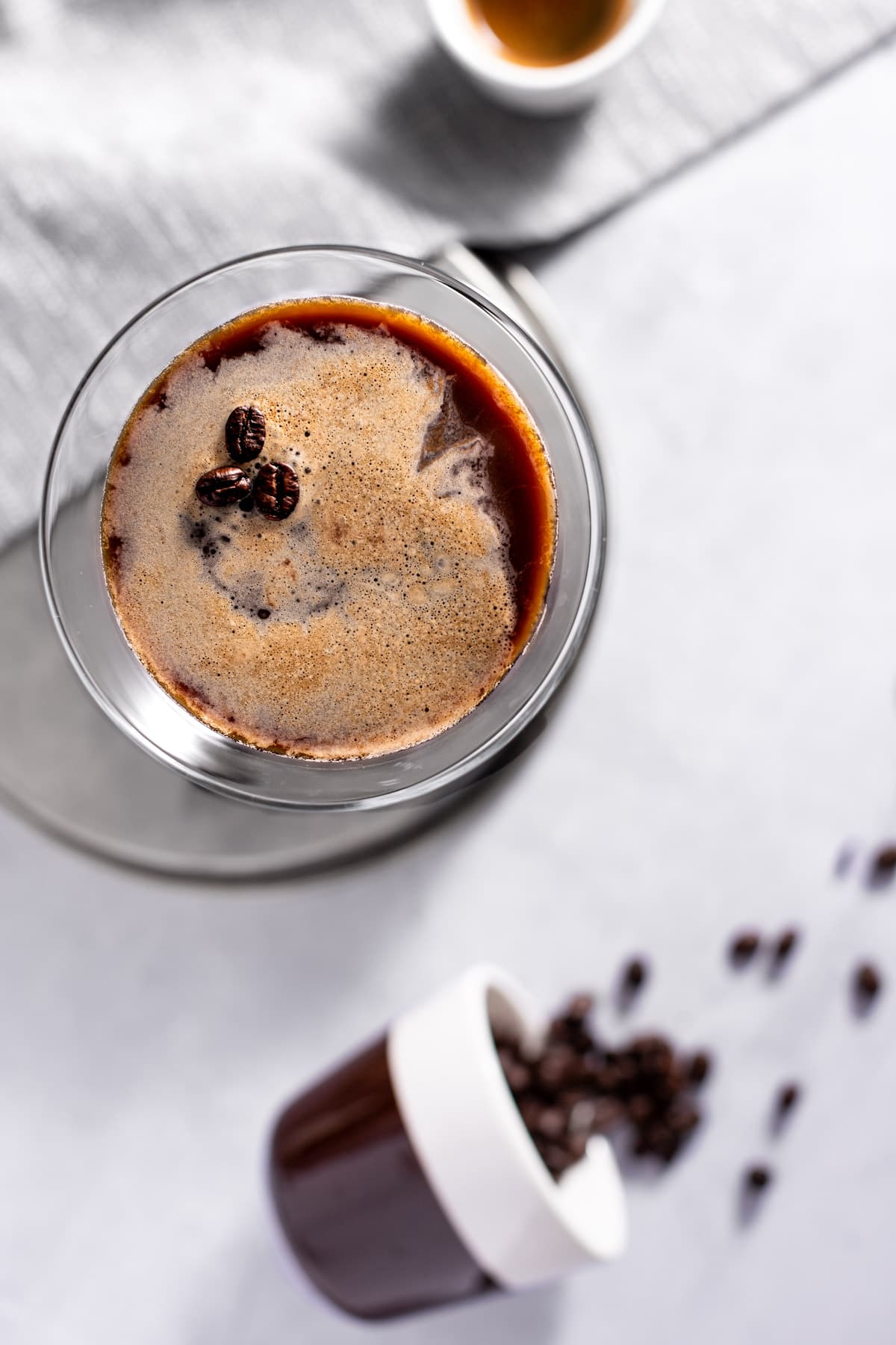 Overhead view of a gin espresso martini, on a grey napkin, next to a cup of tipped over coffee beans.