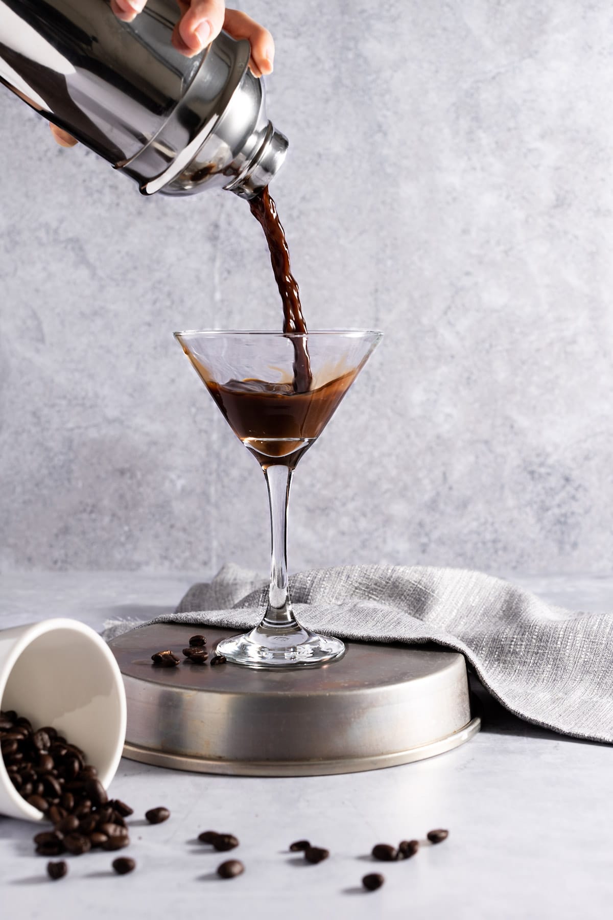 Action shot of the martini being poured from the cocktail shaker into the glass, with spilled coffee beans in front, on a grey background.