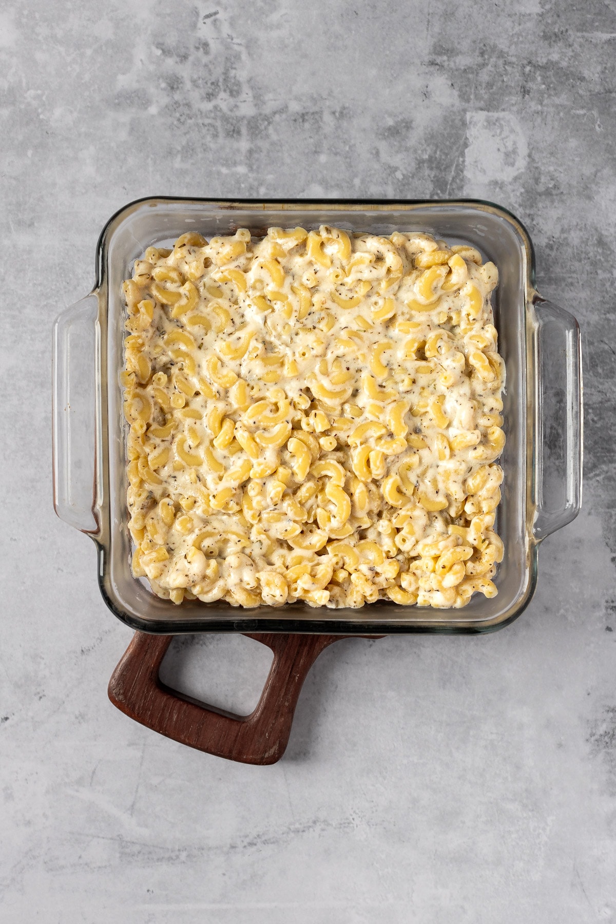 Cooked macaroni pasta that has been coated in the goat cheese sauce, in a square glass baking dish.