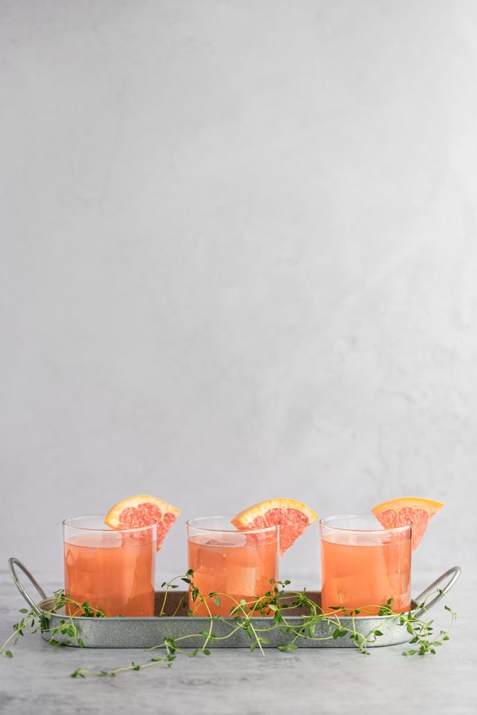 Three grapefruit cocktails beside each other in a metal tray, with fresh thyme scattered at the bottom.