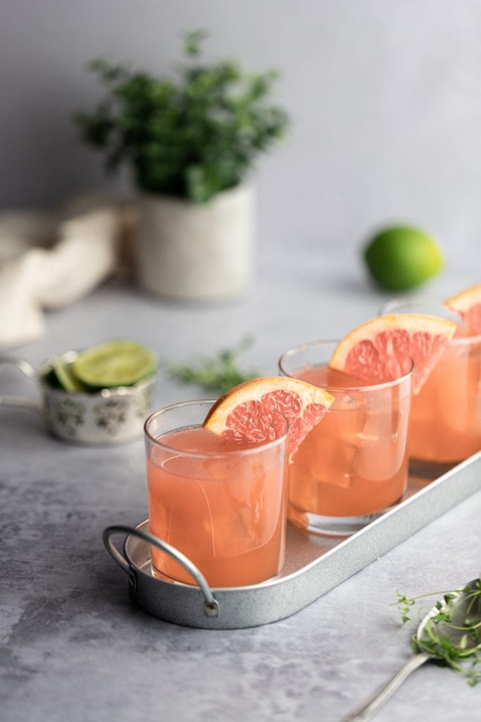 Three grapefruit crush cocktails garnished with grapefruit slices lined up in a rue on an angle in a shiny metal tray, limes in the background.