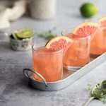 Three grapefruit crush cocktails on a metal serving tray, garnished with grapefruit, limes in the background.