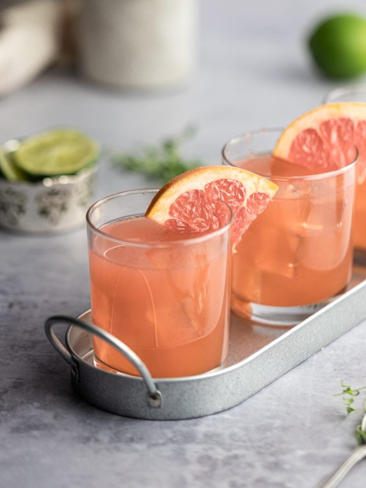 Three grapefruit crush cocktails on a metal serving tray, garnished with grapefruit, limes in the background.