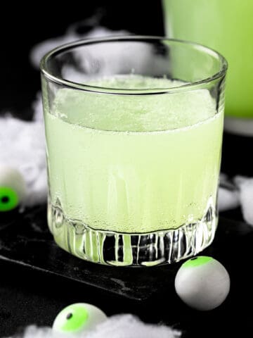 A glass of green halloween punch on a black coaster, next to fake eyeball decorations.