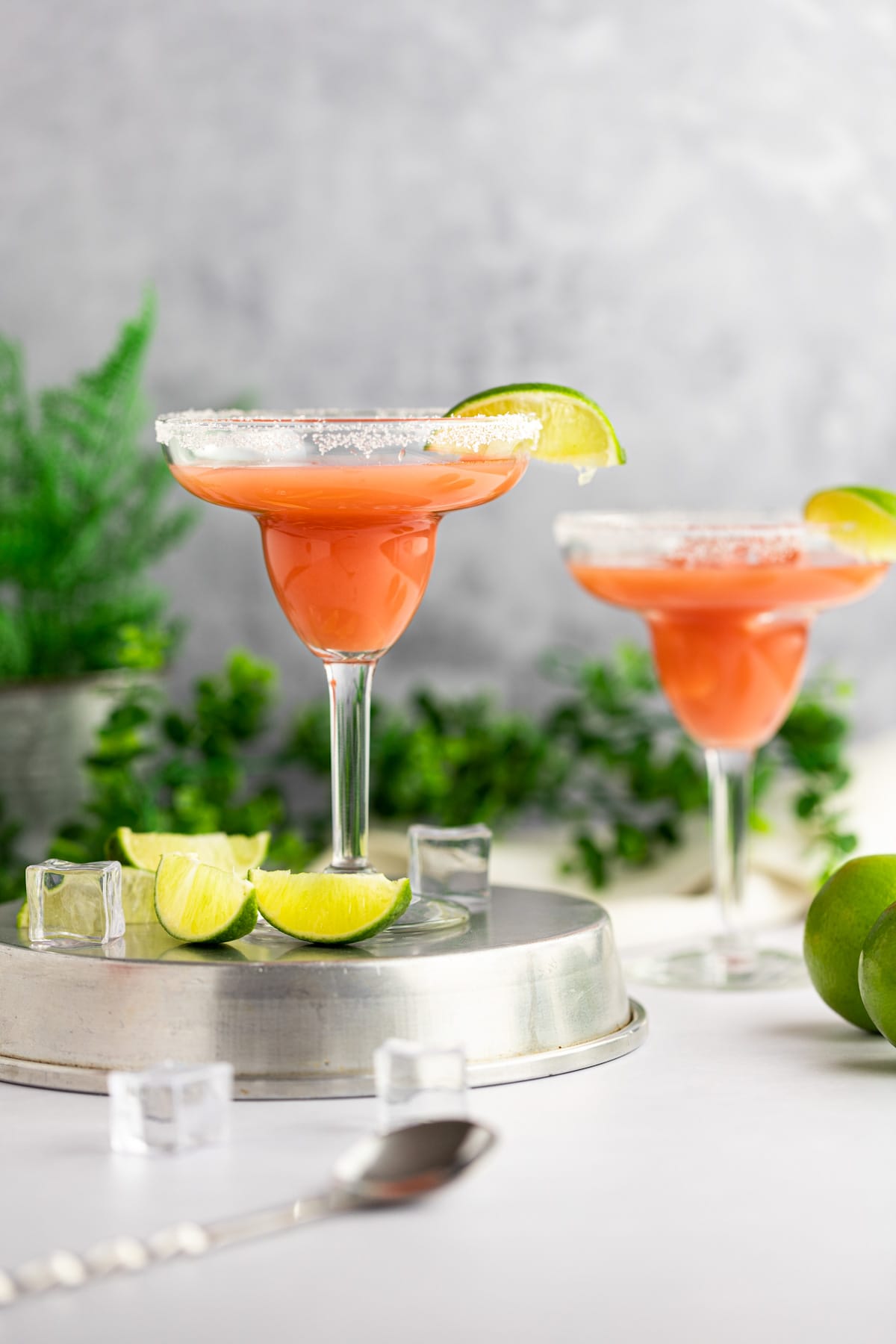 A guava margarita on an upside down round metal tray, next to lime slices and ice cubes, with greenery and another drink in the background.