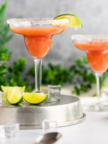 A couple guava margaritas with a slice of lime and salt around the edge of the glass, standing on a metal tin, with green leaves in the background.