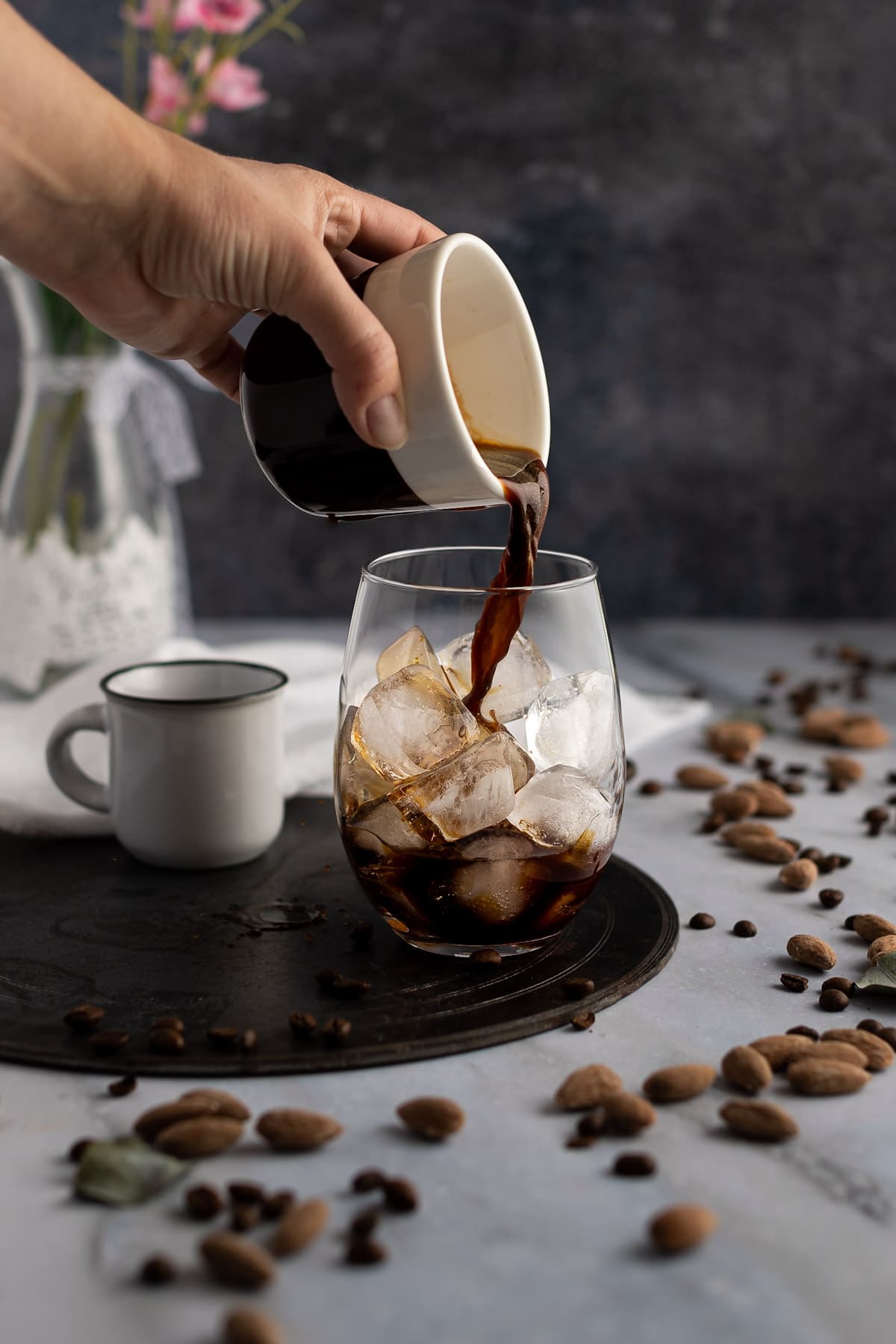A hand pouring a cup of espresso into a glass filled with ice, sitting on a table with scattered almonds and coffee beans.