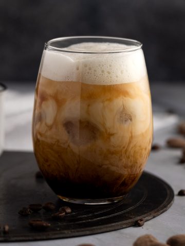 An iced almond milk latte topped with almond milk foam, on a round black metal serving board, with almonds in the background.