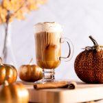 Iced chai latte with pumpkin cold foam on a wooden board, surrounded by decorative pumpkins, and golden leaves in the background.