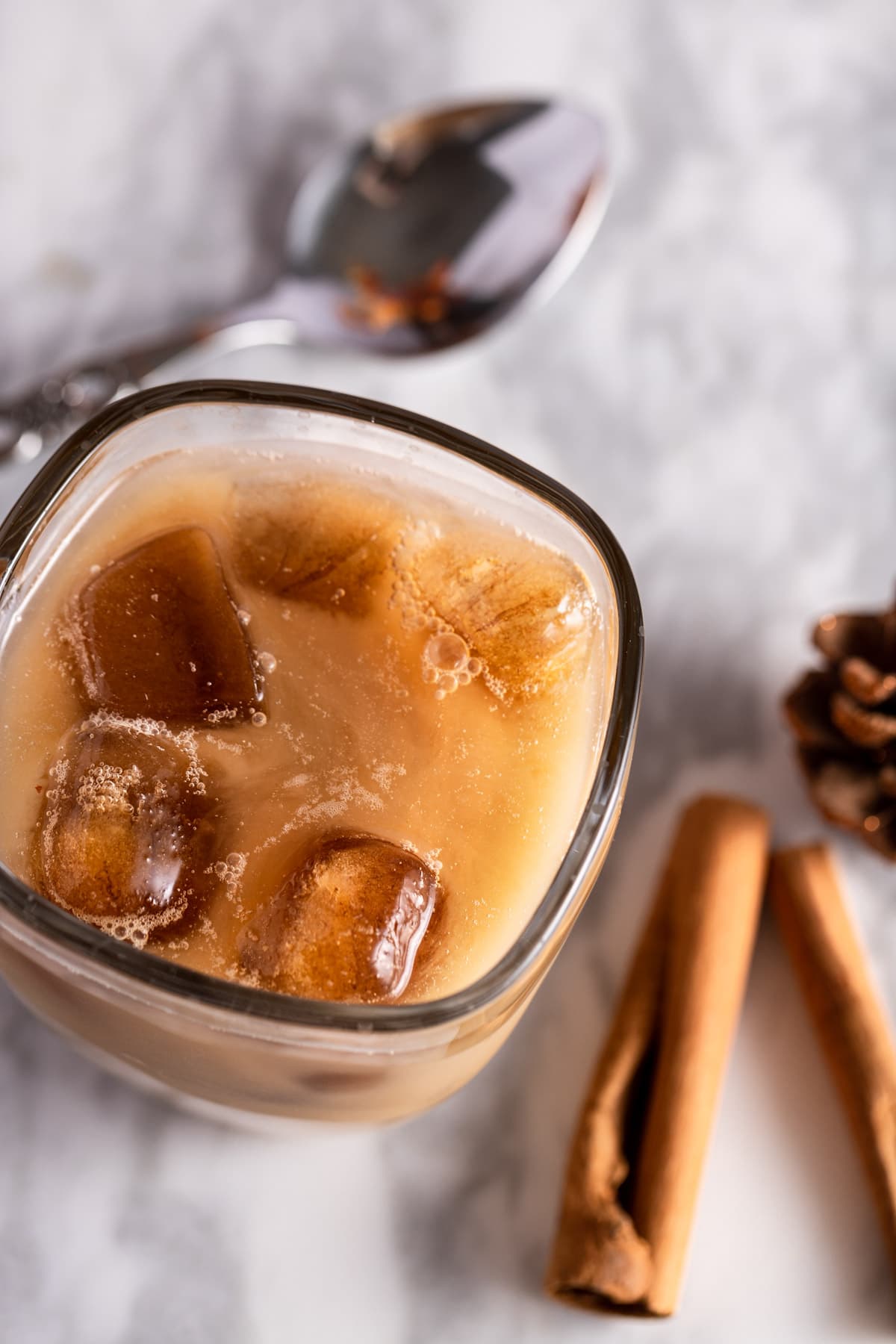Overhead view of an iced chai tea latte with ice cubes, on a marble board, next to cinnamon sticks and a silver spoon.