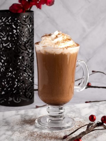 Iced mocha latte with whipped cream and sprinkled with cocoa powder, on a white marble board, surrounded by red berry garlands, and red flowers in the background.