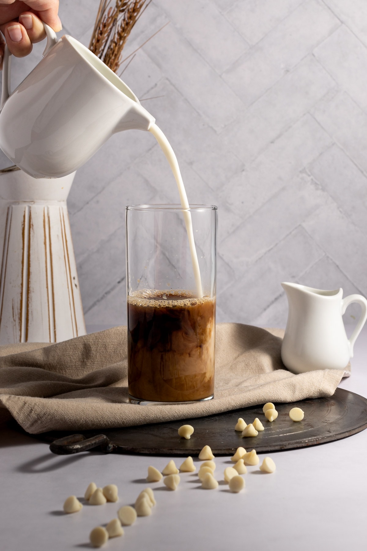 Milk being poured into a tall glass filled with coffee, sitting on a brown napkin, next to scattered white chocolate chips.