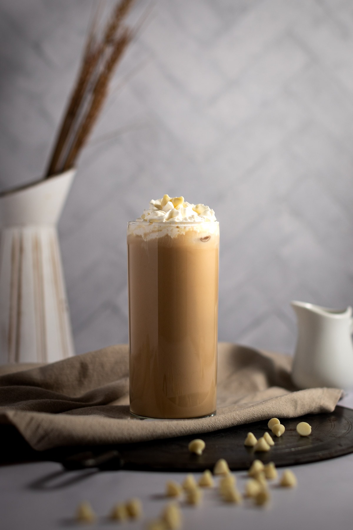 A tall glass of iced white chocolate mocha, topped with whipped cream and white chocolate chips, on a brown napkin and grey background.