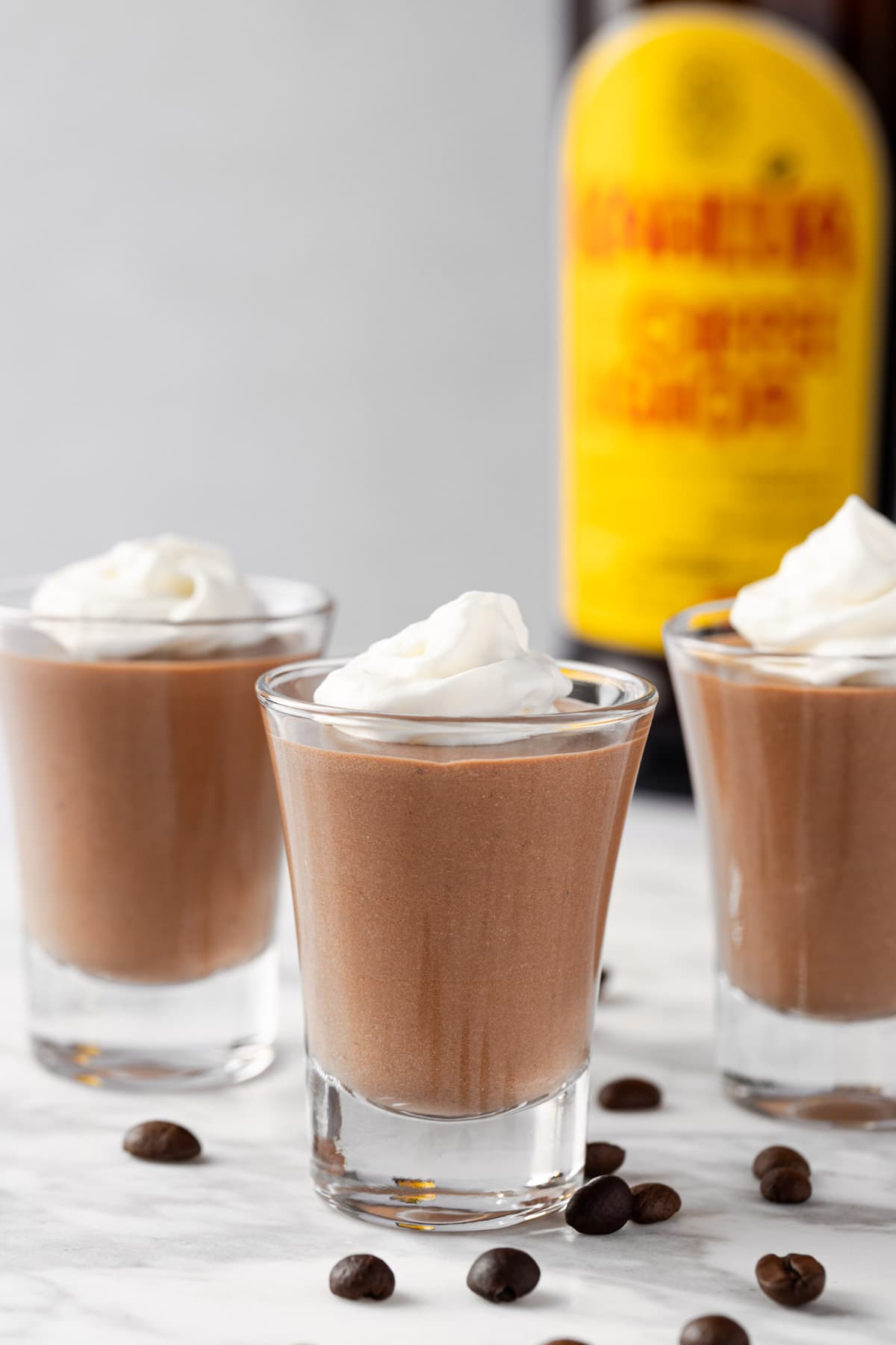 Three Kahlua Pudding Shots topped with whipped cream and a Kahlua bottle in the background.