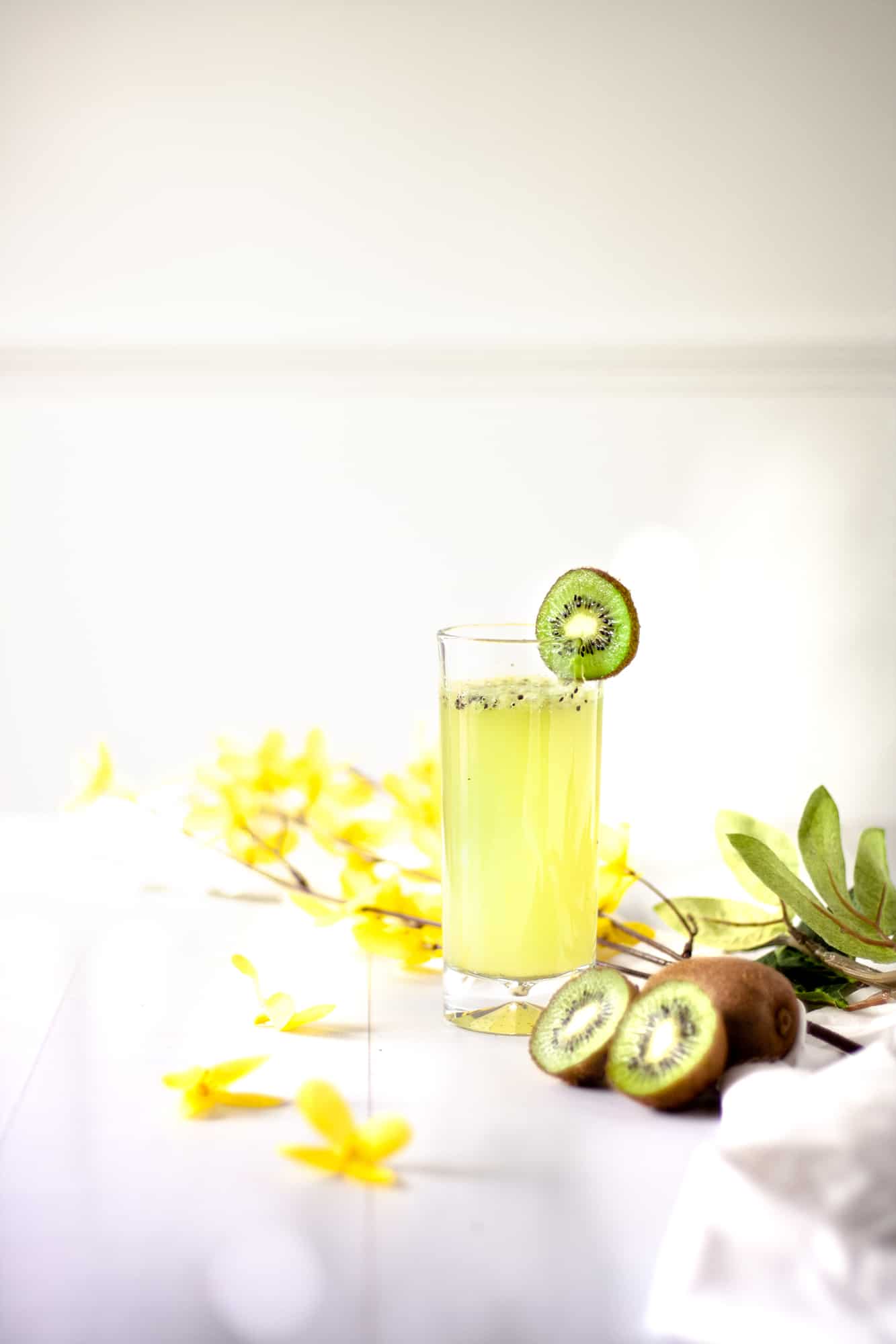 A glass of kiwi gin cocktail, garnished with a slice of kiwi, sitting next to more kiwi slices and a branch of yellow flowers