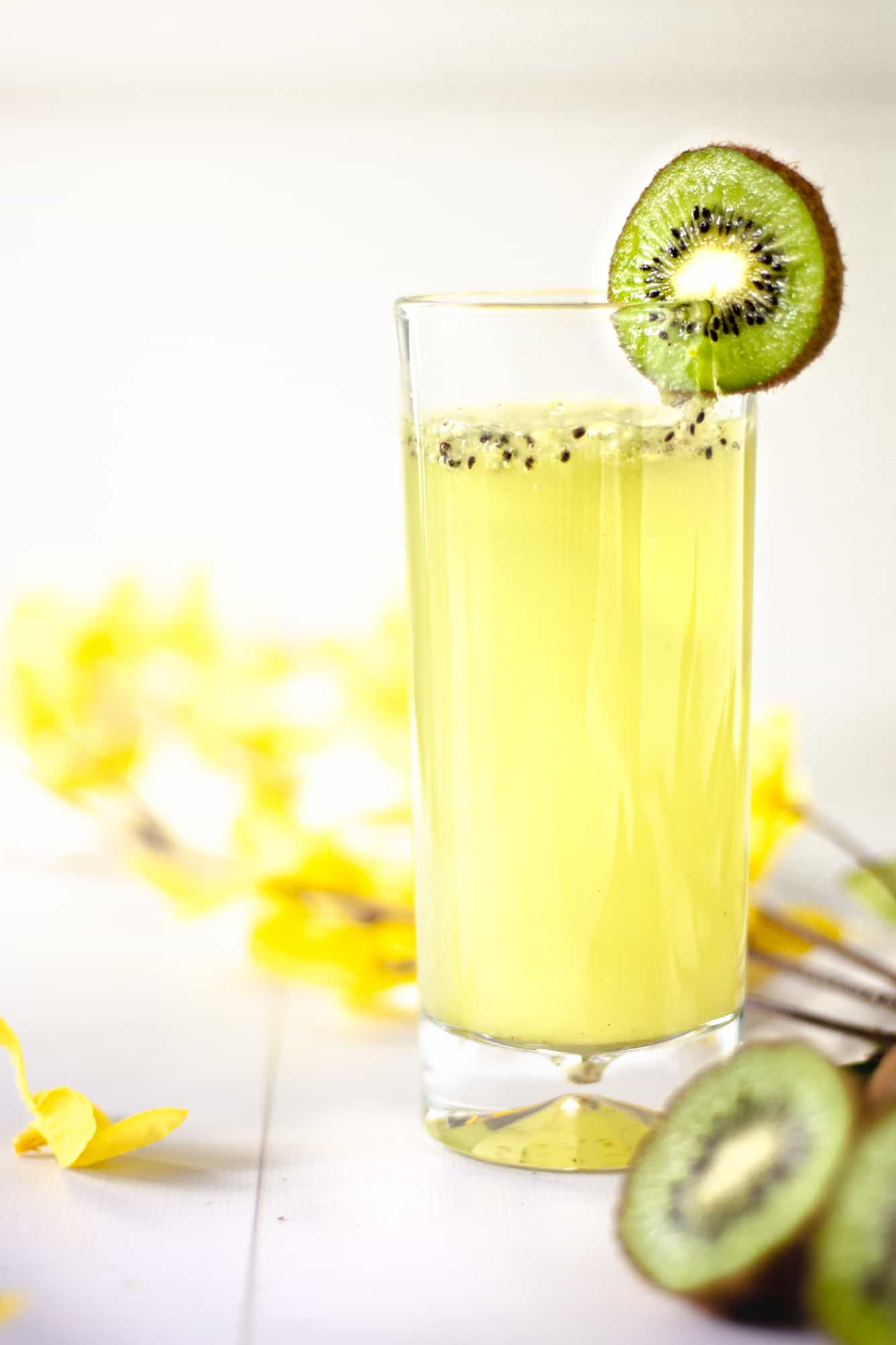 Up close view of a glass of kiwi gin cocktail garnished with kiwi, and kiwis and yellow flowers in the background