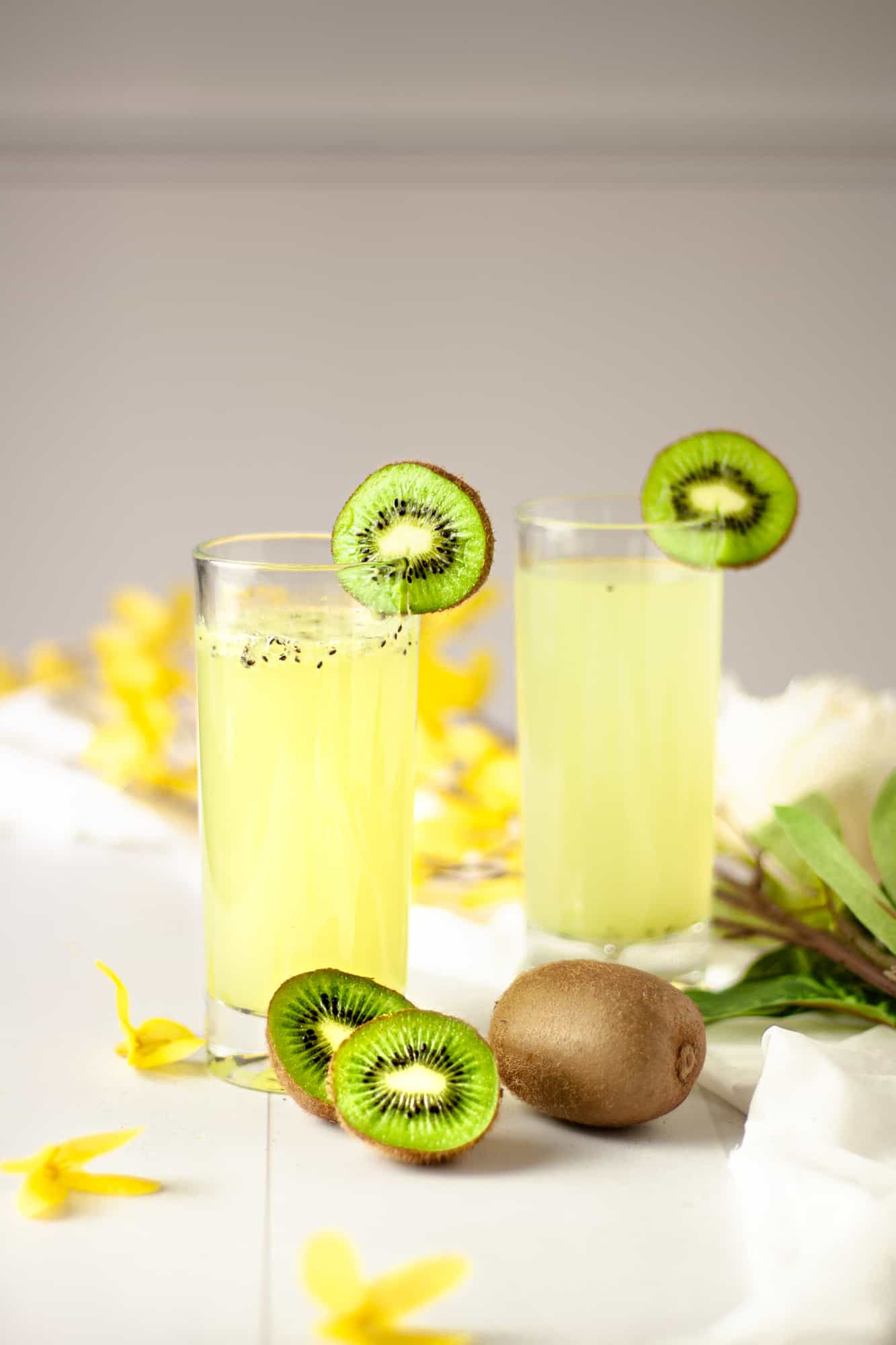 Two kiwi gin cocktails garnished with kiwi slices and sitting in a table with scattered yellow flowers.