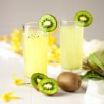 Two kiwi gin cocktails garnished with kiwi slices and sitting in a table with scattered yellow flowers.