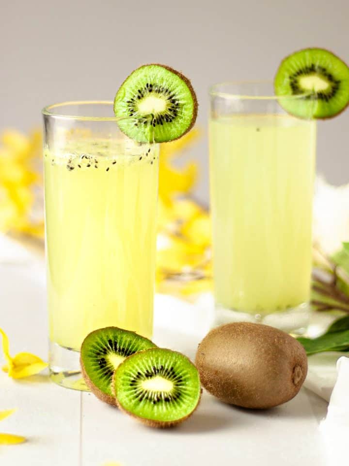 Two kiwi gin cocktails garnished with kiwi slices and sitting in a table with scattered yellow flowers.