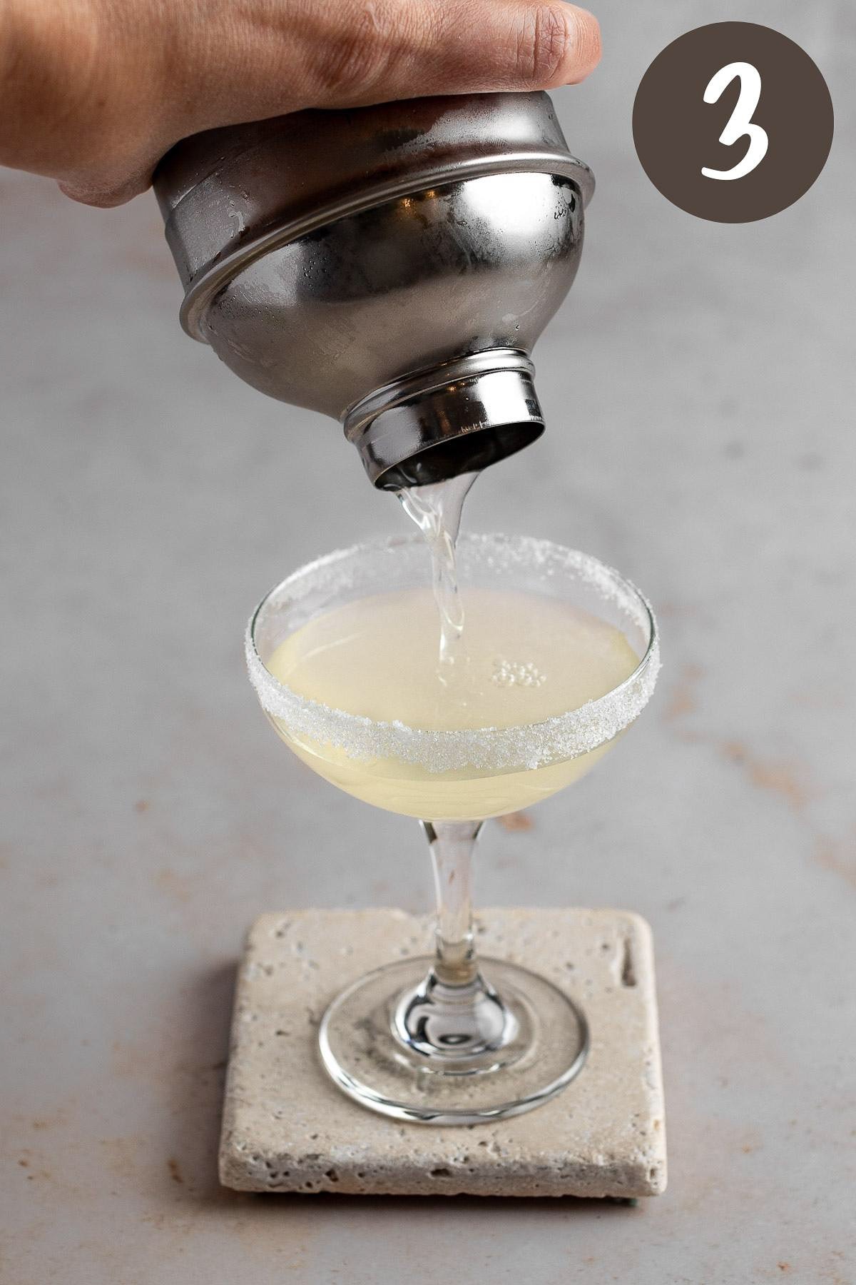 A process photo illustrating the lemon drop martini being poured into a coupe glass.