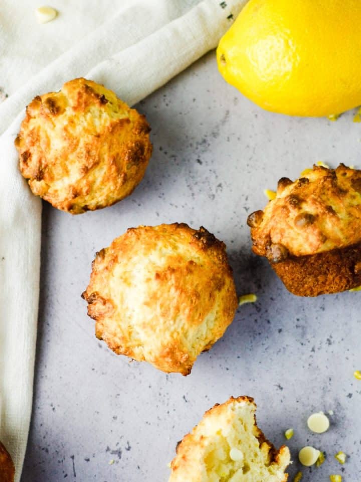 Overhead view of lemon white chocolate muffins scattered on the table with a hand towel, lemon and white chocolate chips