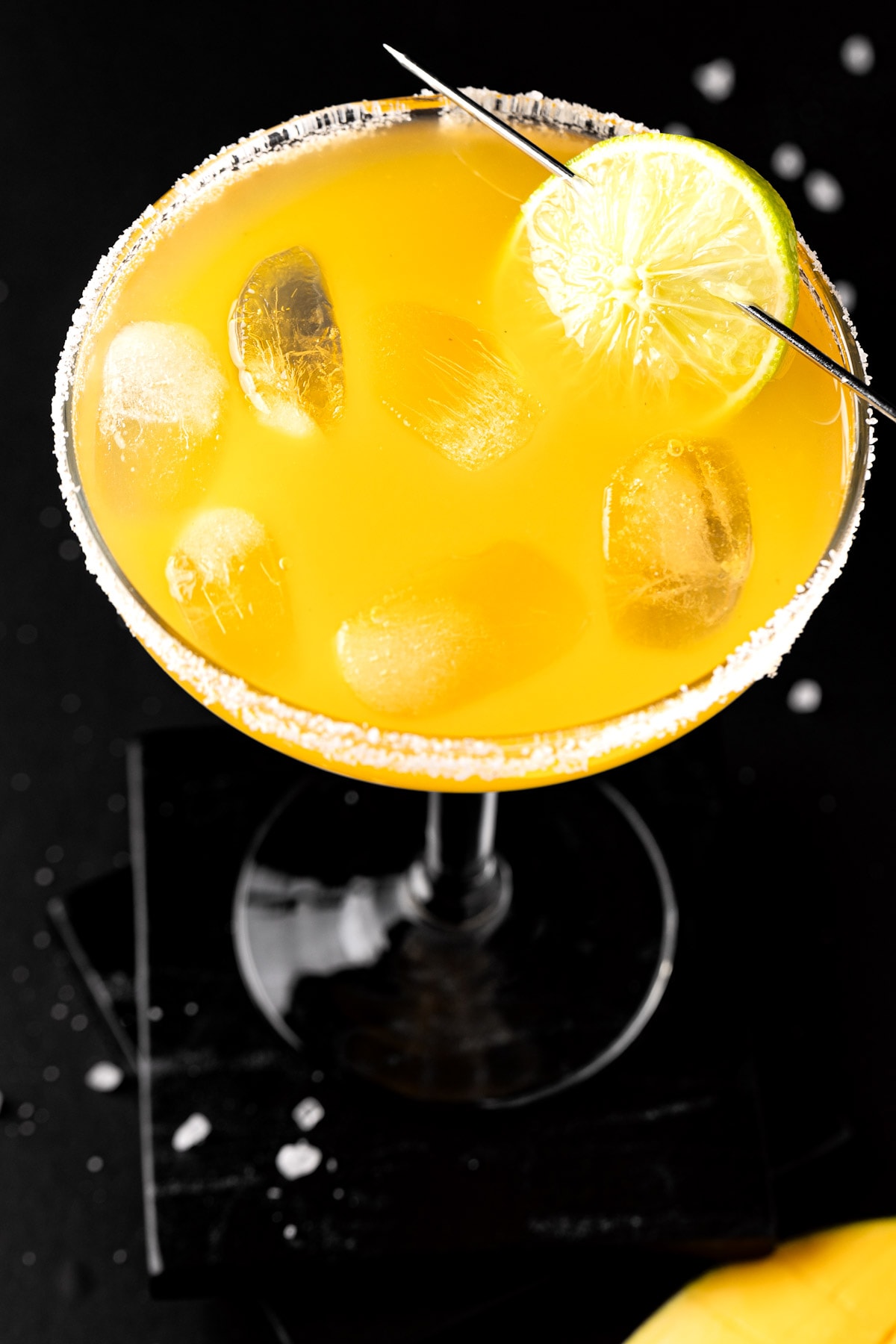 Overhead view of a mango margarita on the rocks, garnished with lime on a black table.