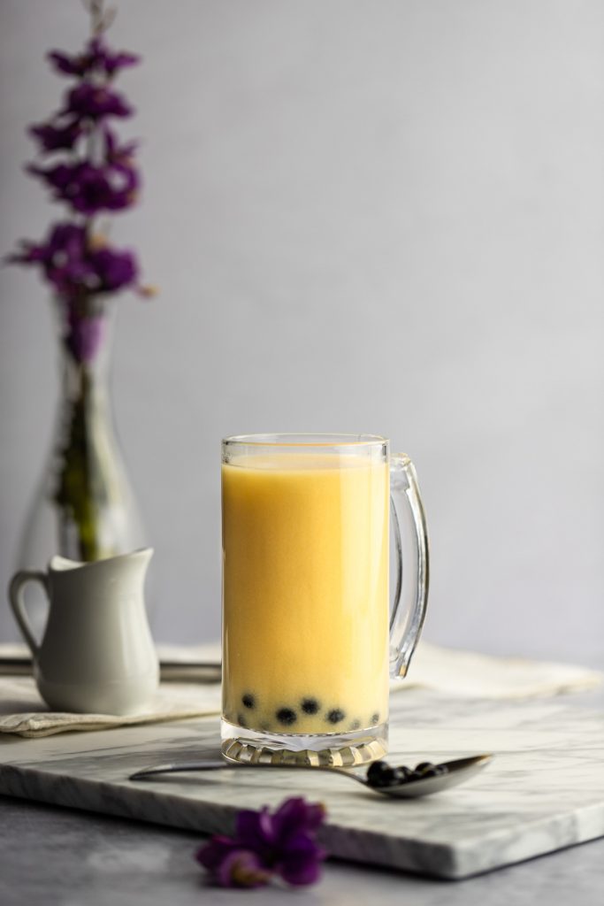 A large yellow mango milk tea on a white marble board, with a spoonful of boba in front and a vase with purple flowers behind.