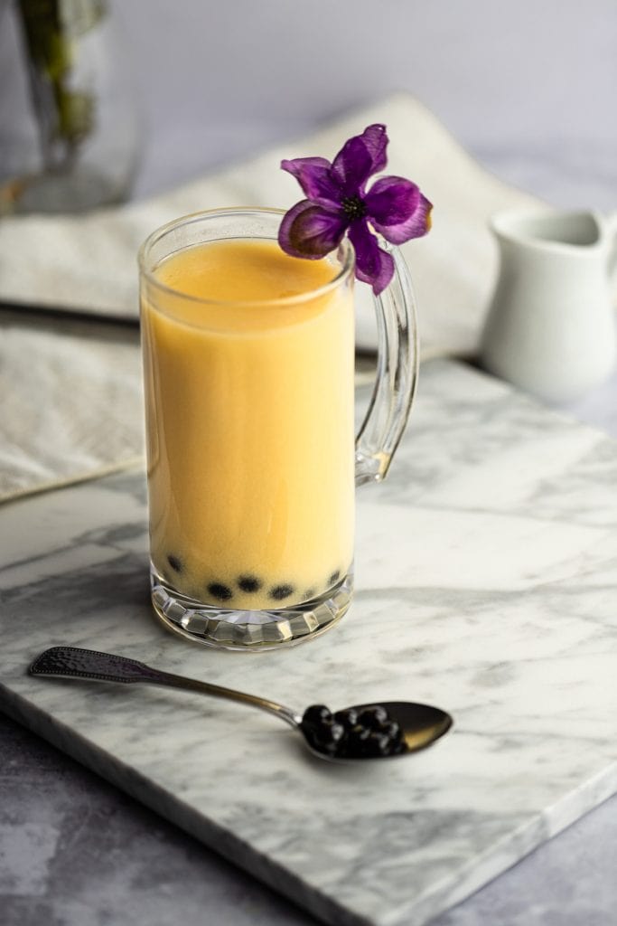 Yellow-orange milk tea with boba on a white marble board, with a spoon of boba, beige napkin in the background.