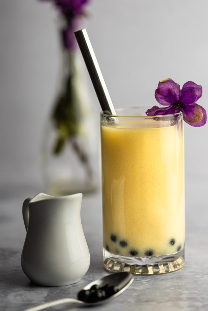 Mango bubble tea with a metal boba straw, purple flower on the rim, next to a small white creamer.