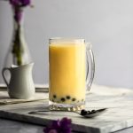 A large mango milk tea with a small creamer and vase of flowers in the background, spoon of boba and purple flower in the foreground.