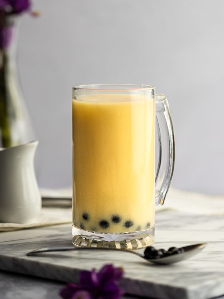 A large mango milk tea with a small creamer and vase of flowers in the background, spoon of boba and purple flower in the foreground.