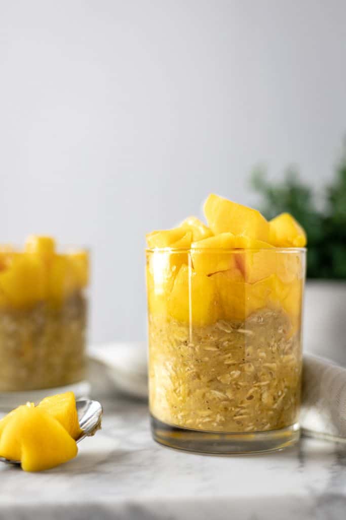 Side angle view of a glass of mango overnight oats, beside a metal spoon full of mango pieces