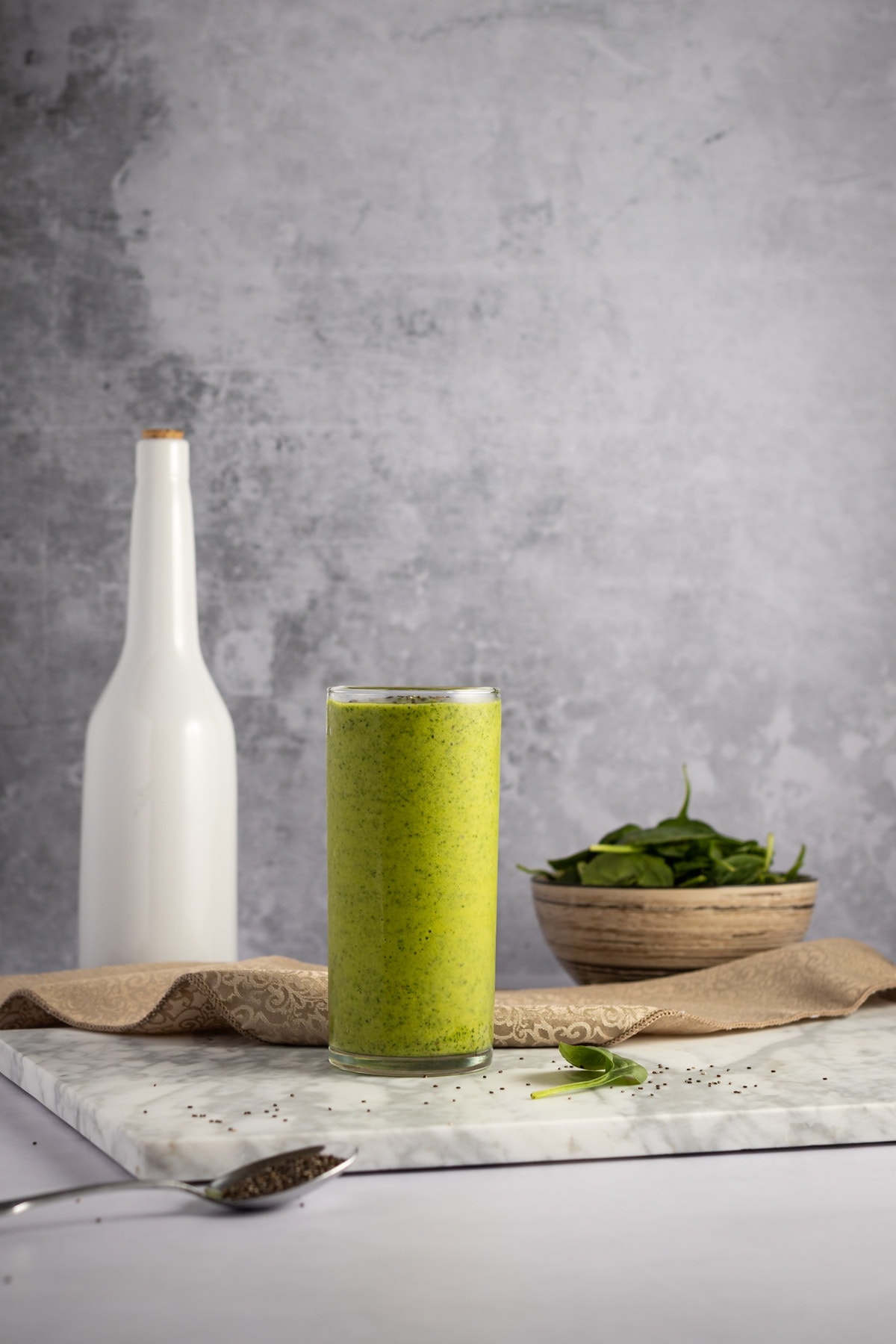 A tall glass of spinach mango smoothie, on a grey marble board, with a tall white jug and brown bowl filled with fresh spinach in the background.