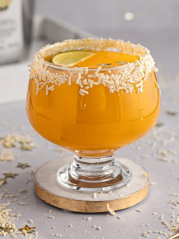 Mango tango drink on with a coconut glass rim, on a white and gold coaster, with gold stars and coconut scattered on the table.