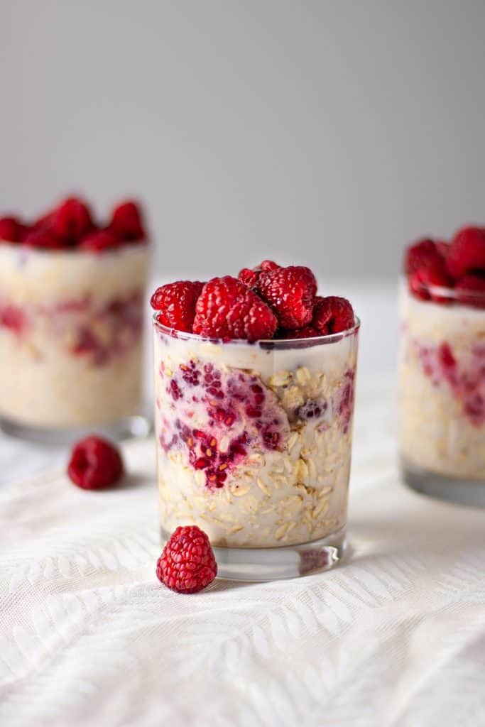 There glasses of maple and raspberry overnight oats with raspberries scattered on the table