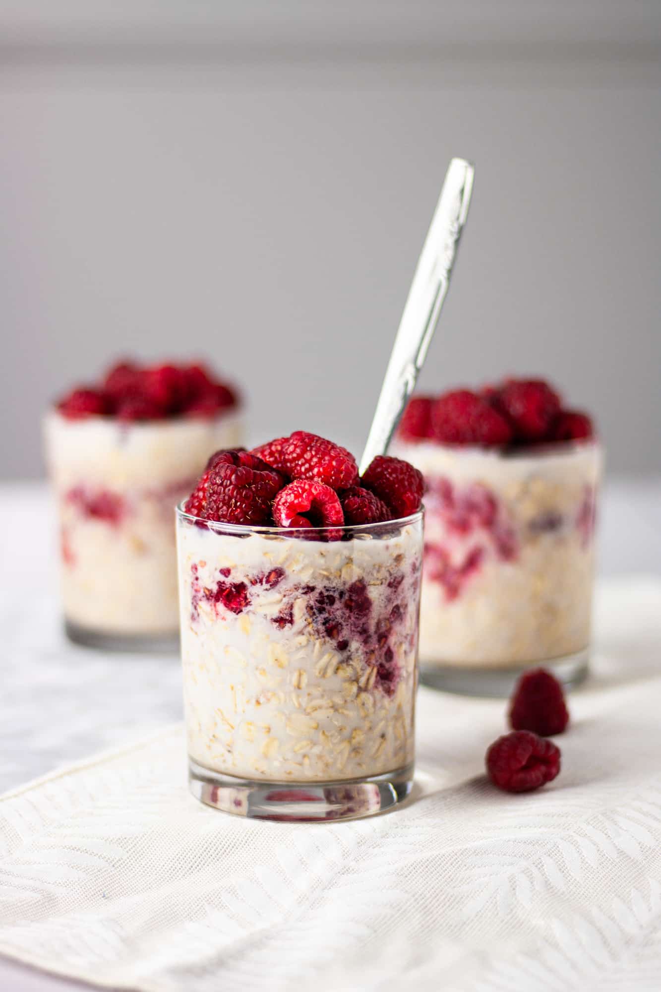 A glass of raspberry maple oats with a silver spoon sticking out of the glass and two other glasses of oats in the background