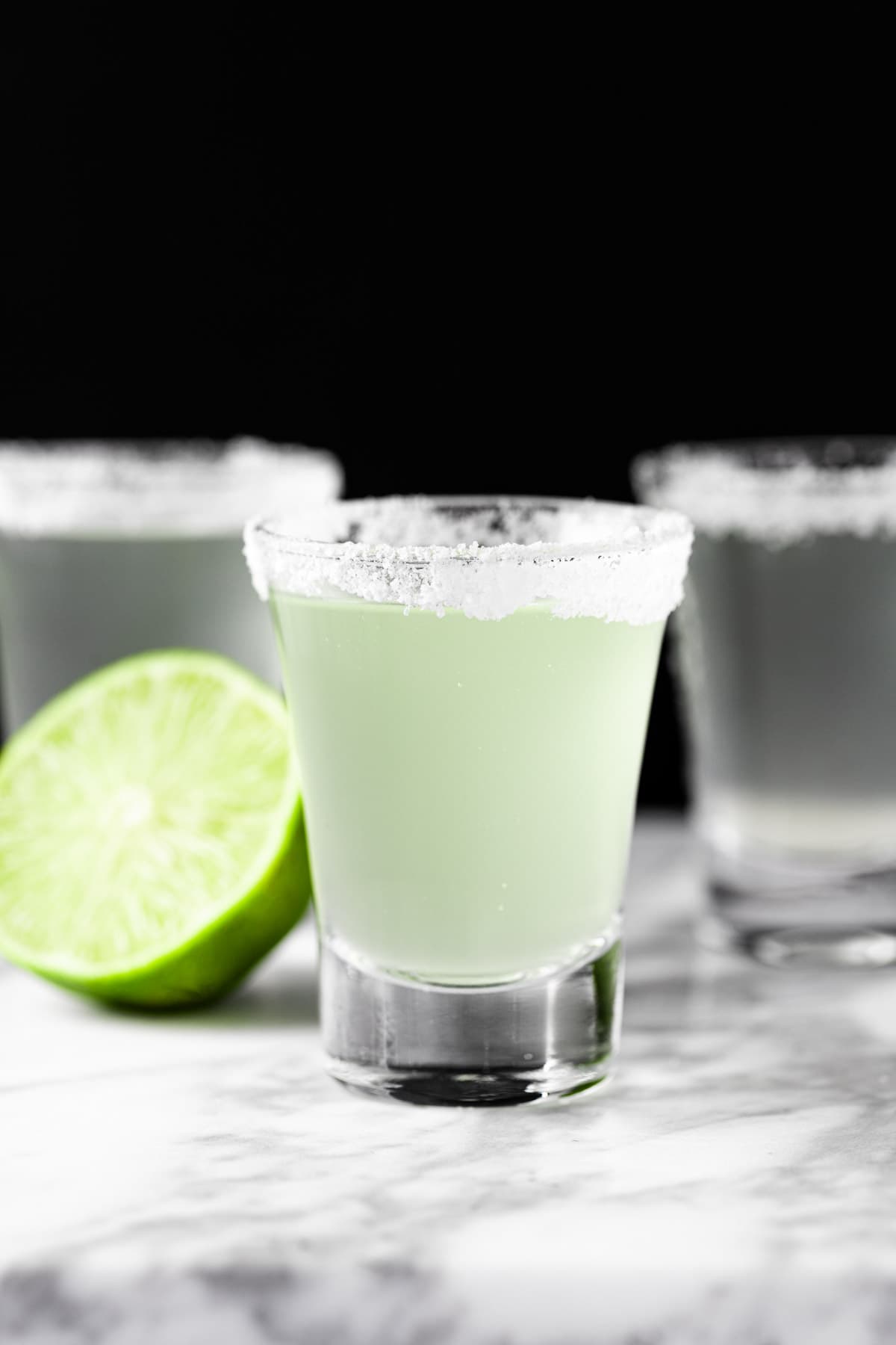 Three margarita shooters with a salted rim, next to half a lime.