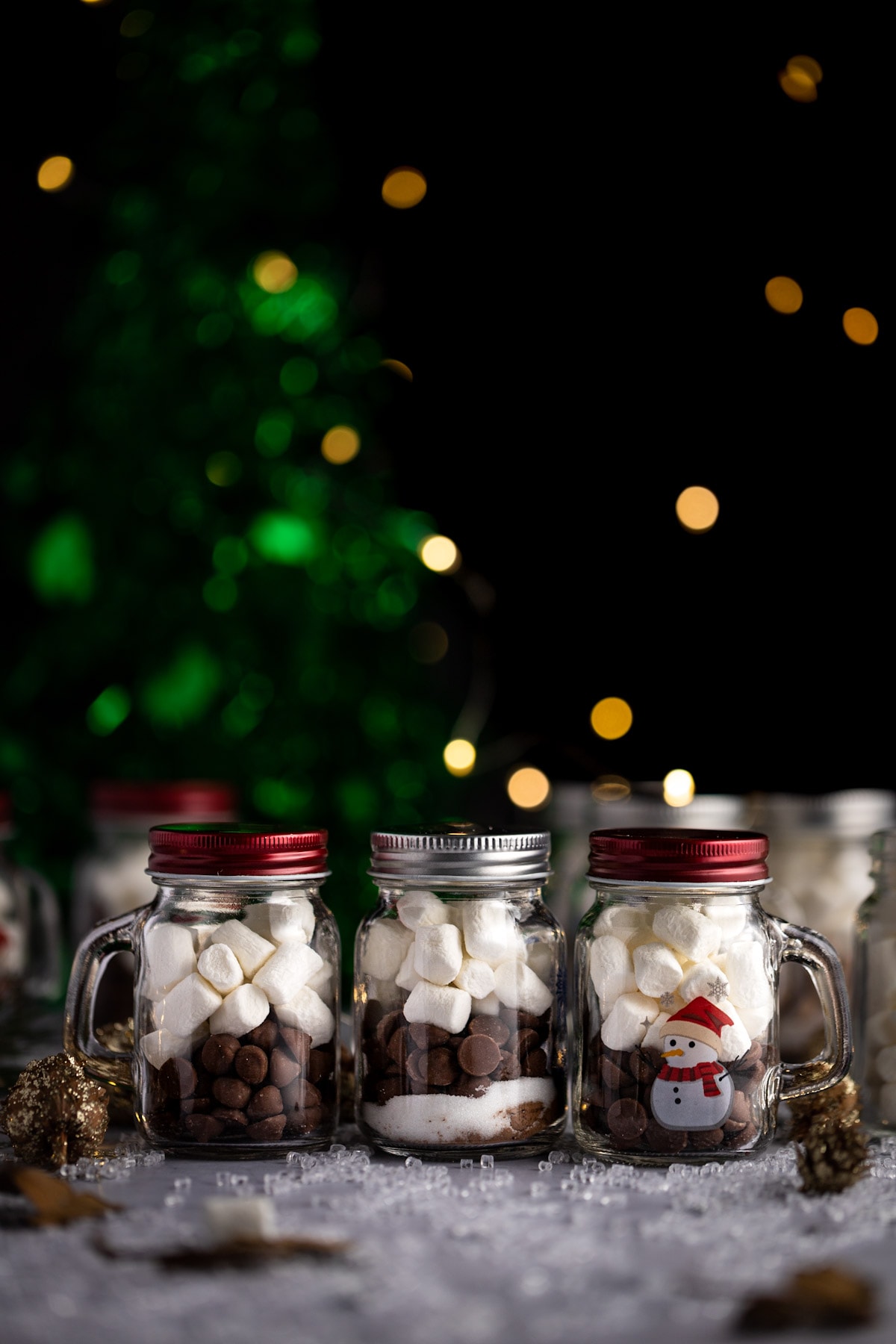 Three small christmas hot chocolate jars in a row, with a christmas tree in the background.