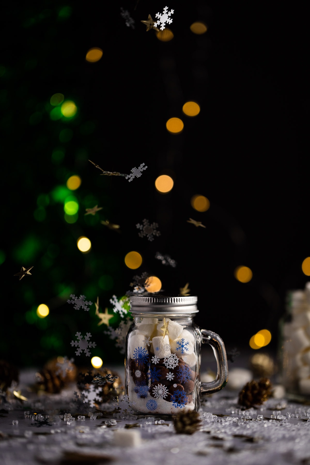Snowflake confetti falling on top of a mini jar filled with hot chocolate mix.