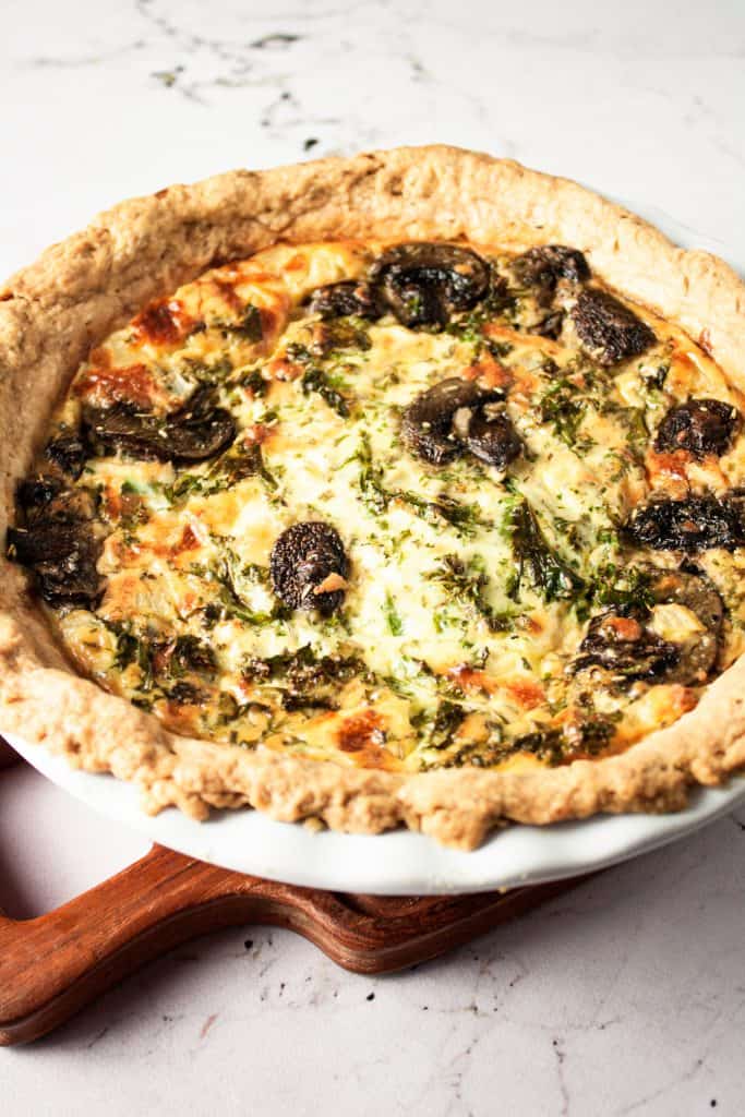 Mushroom kale quiche in a pie dish on a wooden serving board