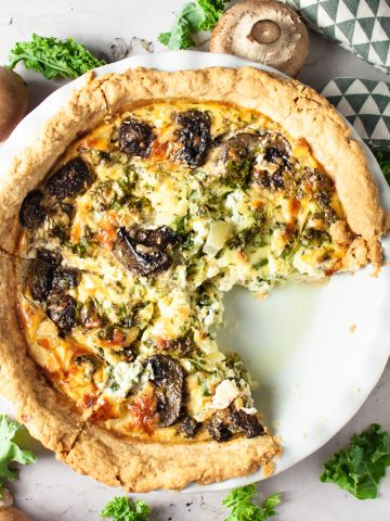 Overhead view of mushroom kale and feta quiche with a slice missing and raw mushrooms and kale spread out on the table