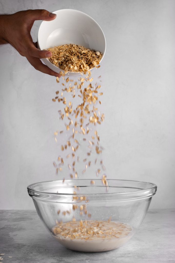 A hand pouring a bowl of overnight oats into a large glass mixing bowl, on a grey background.